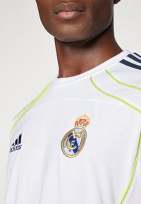 Camisa blanca de Adidas con un logo de Real Madrid en azul y amarillo en el pecho, con acentos verdes y franjas en azul marino en los hombros.