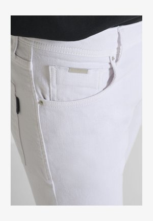 Primer plano de una persona que lleva jeans de mezclilla blancos con un pequeño bolsillo frontal y una blusa negra metida dentro.