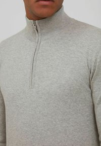 Sudadera de color gris claro con cremallera, cuello acanalado alto, tela texturizada y cremallera de un cuarto en el cuello, con un diseño ajustado.