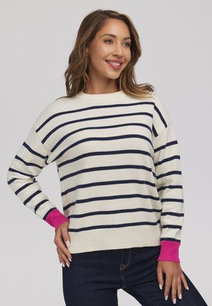 Kobieta uśmiechnięta, ubrana w biały sweter z granatowymi poziomymi paskami i jasnoróżowymi mankietami, w połączeniu z ciemnoniebieskimi dżinsami, na neutralnym tle.