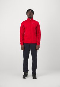 Chaqueta roja con cremallera y cuello alto, con costuras en negro que contrastan; combinada con pantalones azul marino y zapatillas negras con detalles verdes.