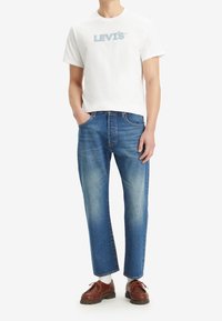 Camiseta blanca de algodón con el logo azul de "LEVI'S", combinada con jeans de pierna recta azul y zapatos de cuero marrón. Corte clásico, estilo casual.