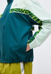 Osoba v tyrkysovo svetlozelenom páperovom kabáte s neónovo zelenými pruhmi s vzorom a žltými nohavicami, rukou vrecku kabáta.