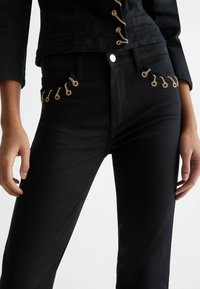 Jeans noirs au design ajusté, ornés d'accents de chaîne dorée le long des poches avant et avec une finition en tissu texturé.