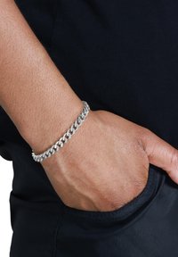 Bracciale in argento con maglie intrecciate. Le maglie testurizzate creano una finitura lucida, indossato su un polso con uno sfondo di tessuto scuro.