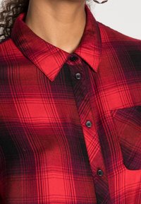 Czerwona i czarna koszula flanelowa w kratę z wyłamiamym kołnierzykiem, z czarnymi guzikami i kieszenią na piersi. Wygląda na miękką i tkana.