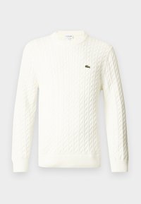 Hvit cable-knit genser med rund hals, lange ermer og en grønn brodert logo på brystet. Laget av et mykt strikket stoff.