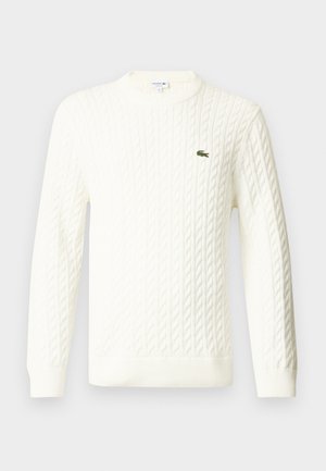 Vit kabelstickad tröja med rund halsringning, långa ärmar och en grön broderad logotyp på bröstet. Tillverkad av ett mjukt stickat tyg.