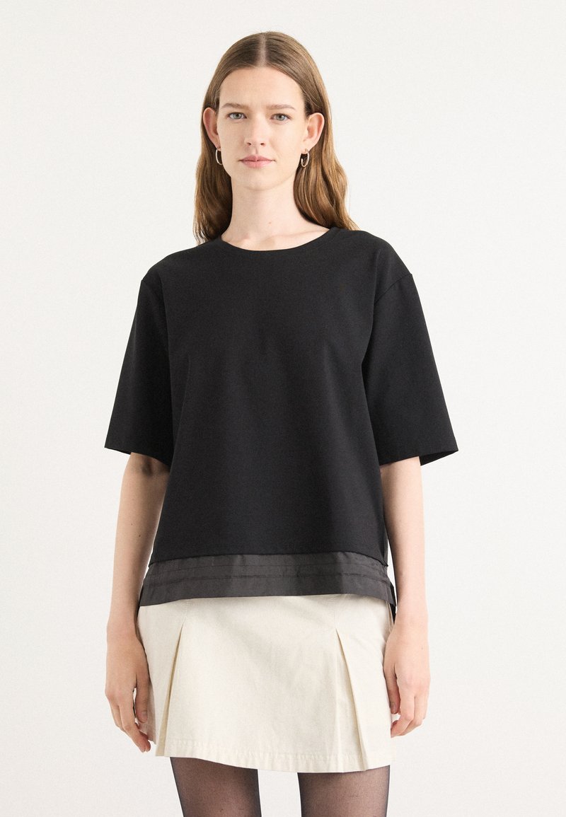 Black cotton t-shirt with short sleeves, round neckline, and a satin band at the bottom, paired with a cream pleated skirt.