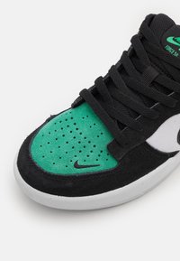 Fekete cipő zöld velúr felsőrésszel, perforálásokkal és fekete fűzőkkel. Fehér középtalppal, amely texturált részleteket tartalmaz. Nike logó az oldalon.