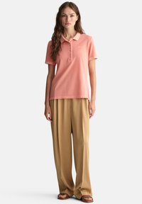 Lyserød polo med korte ærmer, tre knapper foran og subtilt logo. Sammen med beige brede bukser og brune sandaler.