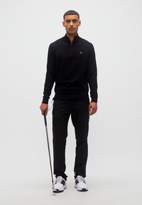 Νεαρός άνδρας που στέκεται με ένα μπαστούνι γκολφ, φορώντας μαύρο μακρυμάνικο μπλουζάκι, μαύρο παντελόνι και λευκά παπούτσια γκολφ σε απλό λευκό φόντο.