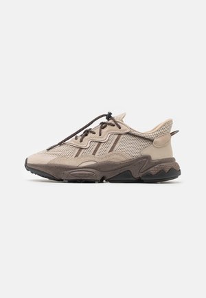 Beige und braune Sportsneaker mit Mesh-Obermaterial, strukturiertem Zwischensohlendesign, gewelltem Seitenmuster und einem mit Zugverschluss befestigten Schnürsystem vor weißem Hintergrund.