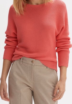 Vrouw draagt een koraalroze ribgebreide trui met opgerolde mouwen en beige broek met hoge taille en knoopsluiting.