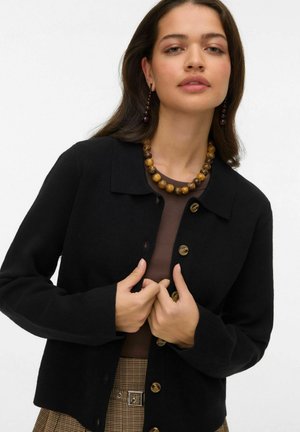 Cardigan noir avec un col à revers, orné de gros boutons marron. Porté sur un haut marron, accessoirisé d'un collier chunky de perles.