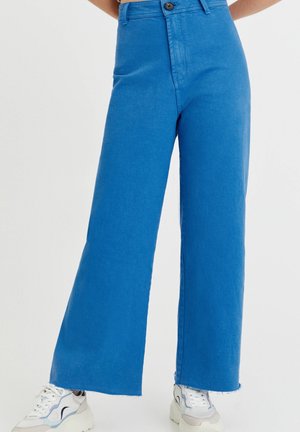 Leg-length view of a person wearing bright blue high-waisted wide-leg pants and white athletic sneakers with gray and blue details.