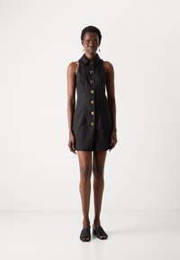 Črna brezrokavna obleka s pasom, ki ima velike zlate gumbe po sprednji strani. Gladka tekstura in prilagojen kroj. Model nosi črne mule.