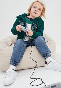 Sudadera con forro de felpa verde, camiseta blanca, pantalones de mezclilla azules y zapatillas blancas. Un niño se sienta en un puff de micropana beige sosteniendo un control de juego.