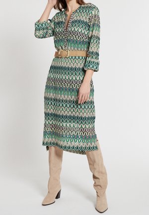Mujer con un vestido estampado ondulado en verde, azul y beige, con mangas remangadas, cinturón beige y botas beige de tacón hasta la rodilla.