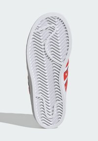 Biely gumový podrážka s textúrovaným zigzagovým vzorom a červenými akcentmi. Obsahuje okrúhle výrezy a vyvýšené okraje pre lepšiu priľnavosť a trvanlivosť.