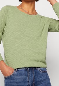Maglione verde chiaro con texture, scollo rotondo e maniche lunghe, caratterizzato da un motivo a nido d'ape. Abbinato a jeans in denim blu.
