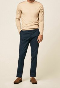 Pull beige à manches longues, pantalon bleu marine et mocassins marron. Le pull a un col rond. Le pantalon est légèrement fuselé. Style minimaliste.