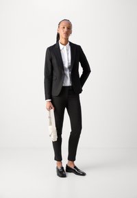 Černý přizpůsobený blazer s klopami, bílá košile s knoflíky, černé slim-fit kalhoty, lesklé �černé loafers a krémová crossbody taška.