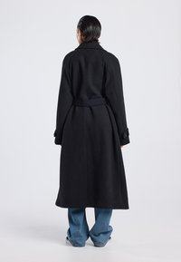 Lange zwarte jas van dik materiaal, met een ceintuur in de taille, brede kraag en knoopsluiting aan de mouwen. Losse pasvorm met een rechte zoom.