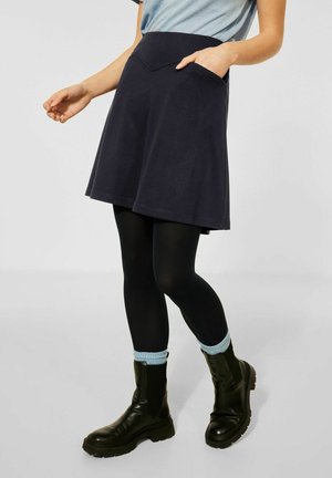 Frau trägt einen marineblauen ausgestellten Rock mit Taschen, schwarze Strumpfhosen, hellblaue Socken und schwarze Stiefeletten, steht vor einem einfarbigen Hintergrund.