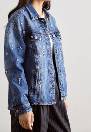 Vrouw draagt een blauwe spijkerjas met kleine geborduurde bloemen, gecombineerd met zwarte pantalon met krijtstrepen en een wit topje.