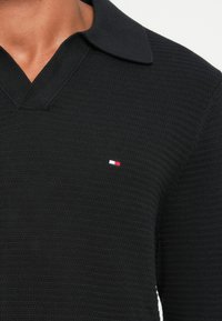 Sort strikket polo skjorte med et tekstureret design og en klassisk krave. Lille logo i rød, hvid og blå på venstre bryst.