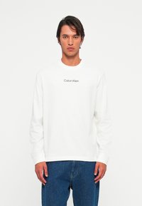 Mann iført hvit langermet Calvin Klein-skjorte og blå jeans, stående mot en ensfarget lys bakgrunn, vendt fremover.