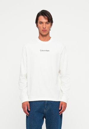 Mann iført hvit langermet Calvin Klein-skjorte og blå jeans, stående mot en ensfarget lys bakgrunn, vendt fremover.