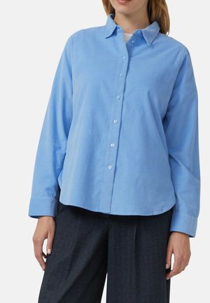 Kobieta ubrana w jasnoniebieską koszulę na guziki z długimi rękawami oraz ciemnoszare spodnie w prążki na białym tle.
