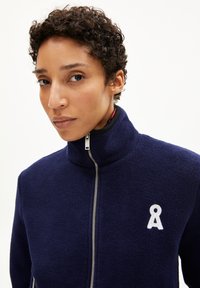 Veste en polaire marine avec un col montant, fermeture éclair argentée et logo brodé blanc sur la poitrine. Fabriquée dans un tissu texturé et doux.