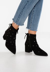 Persona che indossa jeans azzurri con orlo sfrangiato e stivaletti neri in suede con tacco spesso e decorazioni a borchie metalliche.