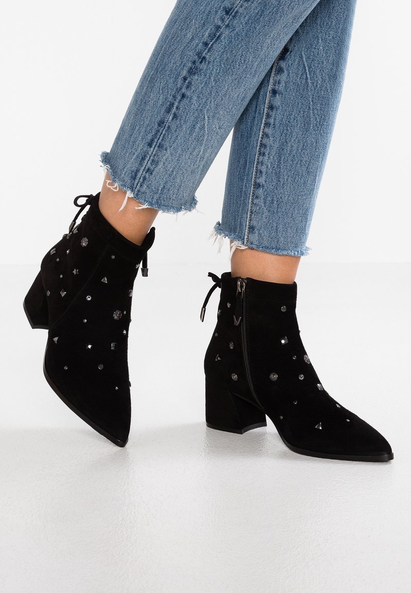 Persona che indossa jeans azzurri con orlo sfrangiato e stivaletti neri in suede con tacco spesso e decorazioni a borchie metalliche.