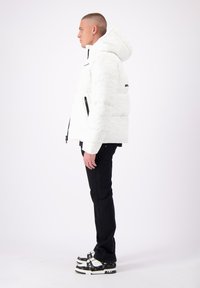 Witte pufferjack met een matte afwerking, capuchon en zwarte ritszakken aan de zijkanten. Zwarte broek en met patroon versierde sneakers maken de outfit compleet.