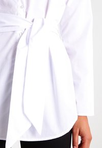 White button-up shirt with a tied waist. Features a smooth, lightweight fabric and long sleeves with a rounded hem.