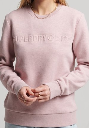 Kvinde iført lyserød Superdry-sweatshirt med præget logo, guldkædehalskæde og ringe på fingrene, stående foran en ensfarvet baggrund.