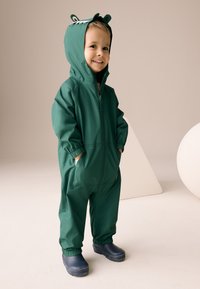 Grøn vandtæt heldragt med lynlåslukning, sidelommer og hætte med dyreagtige ører og øjne. Kombineret med mørkeblå støvler.