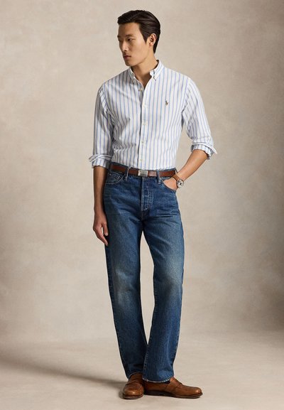Mann trägt ein blau-weiß gestreiftes Hemd mit Knöpfen, blaue Jeans, braunen Gürtel, Uhr und braune Lederschuhe und steht mit einer Hand in der Hosentasche.