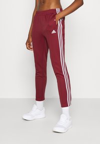 Bordeauxfarbene Sporthose mit schmalem Schnitt, weißen Seitenstreifen und Druckknöpfen entlang der äußeren Nähte. Kombiniert mit weißen Sneakern.
