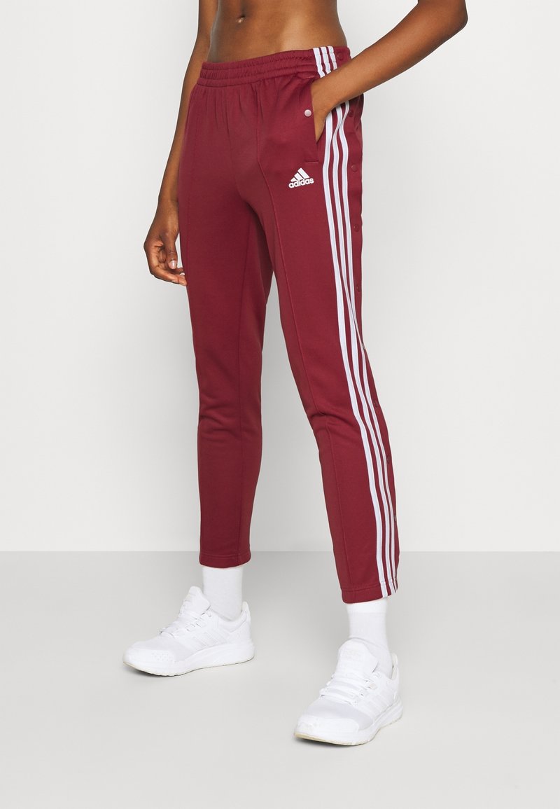 Bordeauxfarbene Sporthose mit schmalem Schnitt, weißen Seitenstreifen und Druckknöpfen entlang der äußeren Nähte. Kombiniert mit weißen Sneakern.