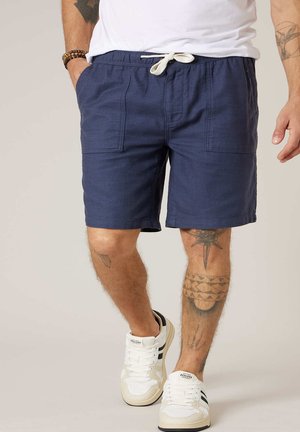 Homme portant un short à cordon bleu marine, des baskets blanches et une chemise blanche, montrant des tatouages sur ses jambes et ses bras.