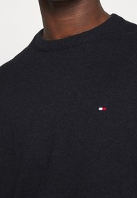 Primo piano di una persona che indossa un maglione a collo rotondo nero in maglia, con un piccolo logo rettangolare rosso, bianco e blu sul petto.