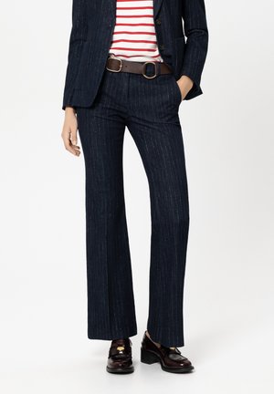 Woman wearing navy pinstripe suit pants and jacket with brown belt, red and white striped shirt, and dark brown heeled loafers.