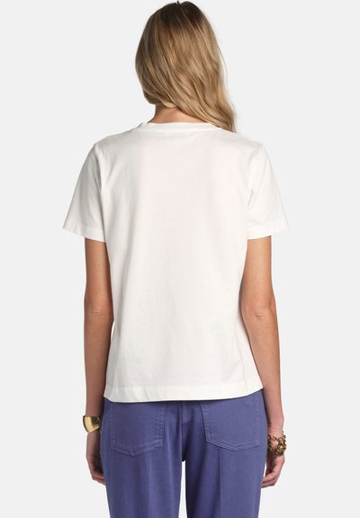 Hátulnézetből egy hosszú szőke hajú nő, egyszerű fehér rövid ujjú ingben és kék nadrágban, egyszínű háttér előtt.