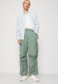 Lyseblå og hvide stribede skjorte med knapper, hvid t-shirt under, grønne cargo-bukser og cremefarvede sneakers. Casual og afslappet pasform.