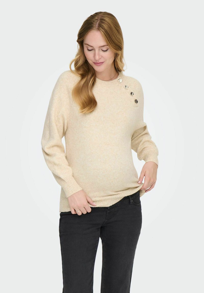 Maglione beige a maglia con maniche lunghe, polsini a costine e bottoni decorativi sulla spalla sinistra. Abbinato a jeans neri. Texture morbida.